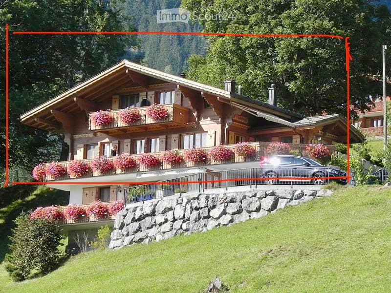 A large, multi-story wooden house with balconies adorned with pink flowers, situated on a hillside with a scenic mountain view in the background.