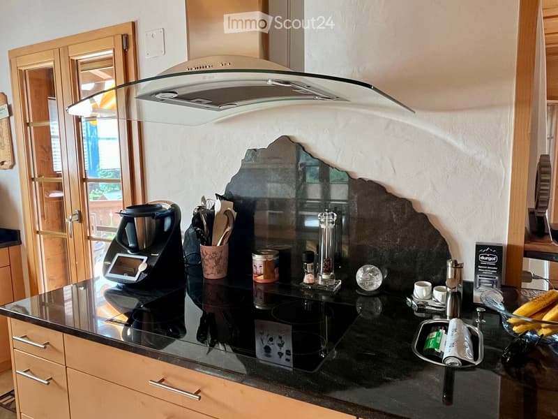 Modern kitchen with a black countertop, induction stove, coffee maker, and various kitchen utensils. The backsplash is dark with a unique, textured design. Wooden cabinets and a glass range hood are visible.