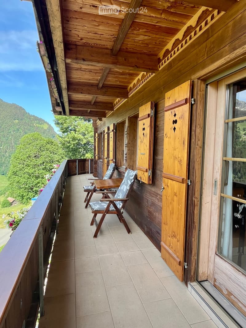 Wooden balcony with tiled floor, two wooden chairs, small table, scenic mountain view.