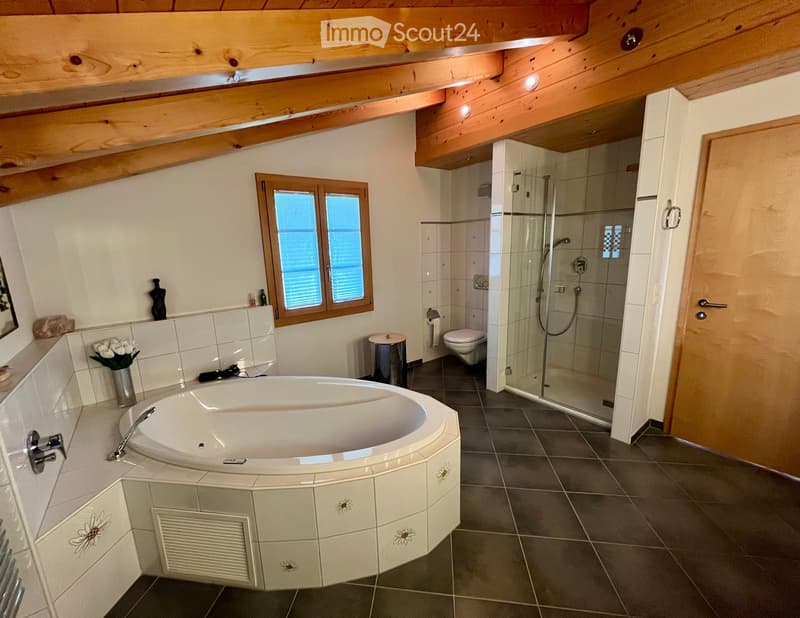 Modern bathroom with a large bathtub, separate shower, toilet, and sink. Features exposed wooden beams on the ceiling, a window with blinds, and decorative elements including a vase with flowers and a small table.