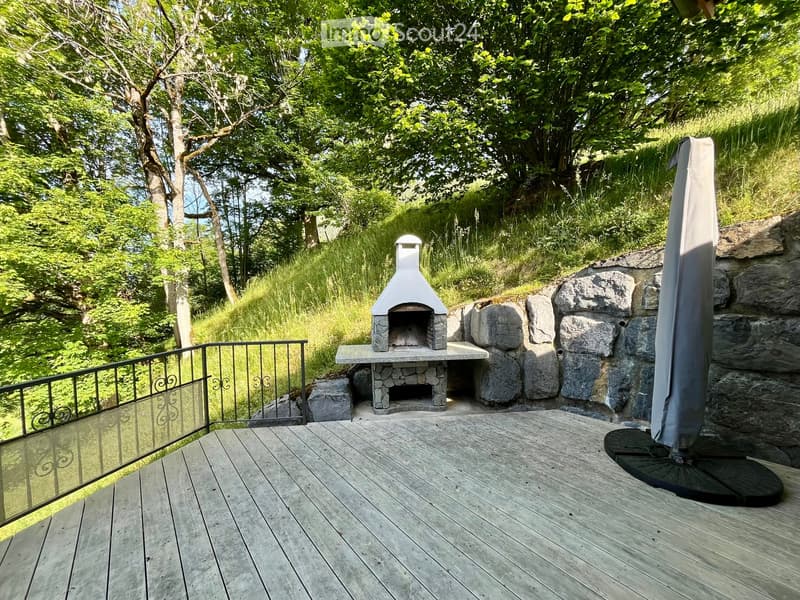 Wooden deck with a stone pizza oven, surrounded by lush greenery and trees, with a metal railing and a covered umbrella stand.