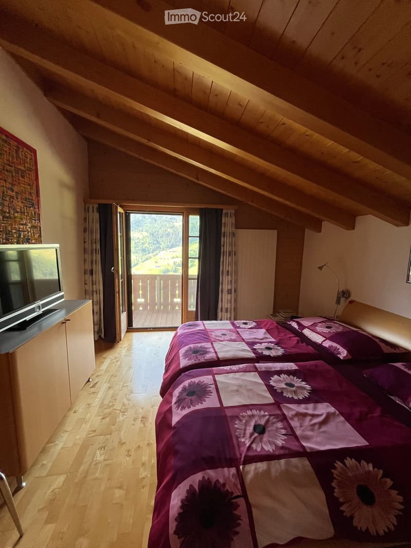 Cozy attic bedroom with wooden slanted ceilings, two single beds with floral bedding, a television on a stand, and a large window offering a scenic view of the surrounding landscape.