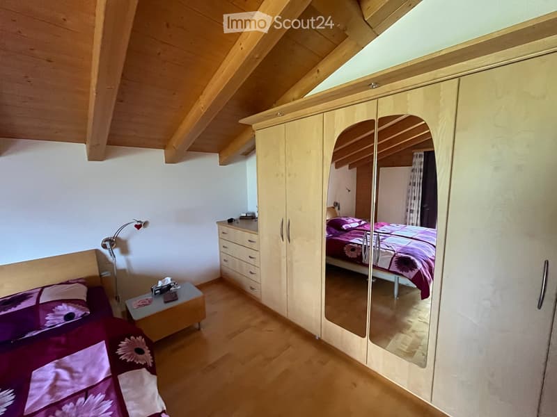 Cozy attic bedroom with wooden slanted ceilings, a large mirrored wardrobe, a bed with purple bedding, a nightstand with a lamp, and a small table with a phone and other items.