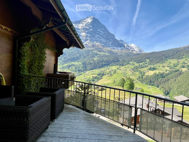 Balcony with wooden decking, black metal railing, and wicker chairs, overlooking a scenic valley and snow-capped mountains.