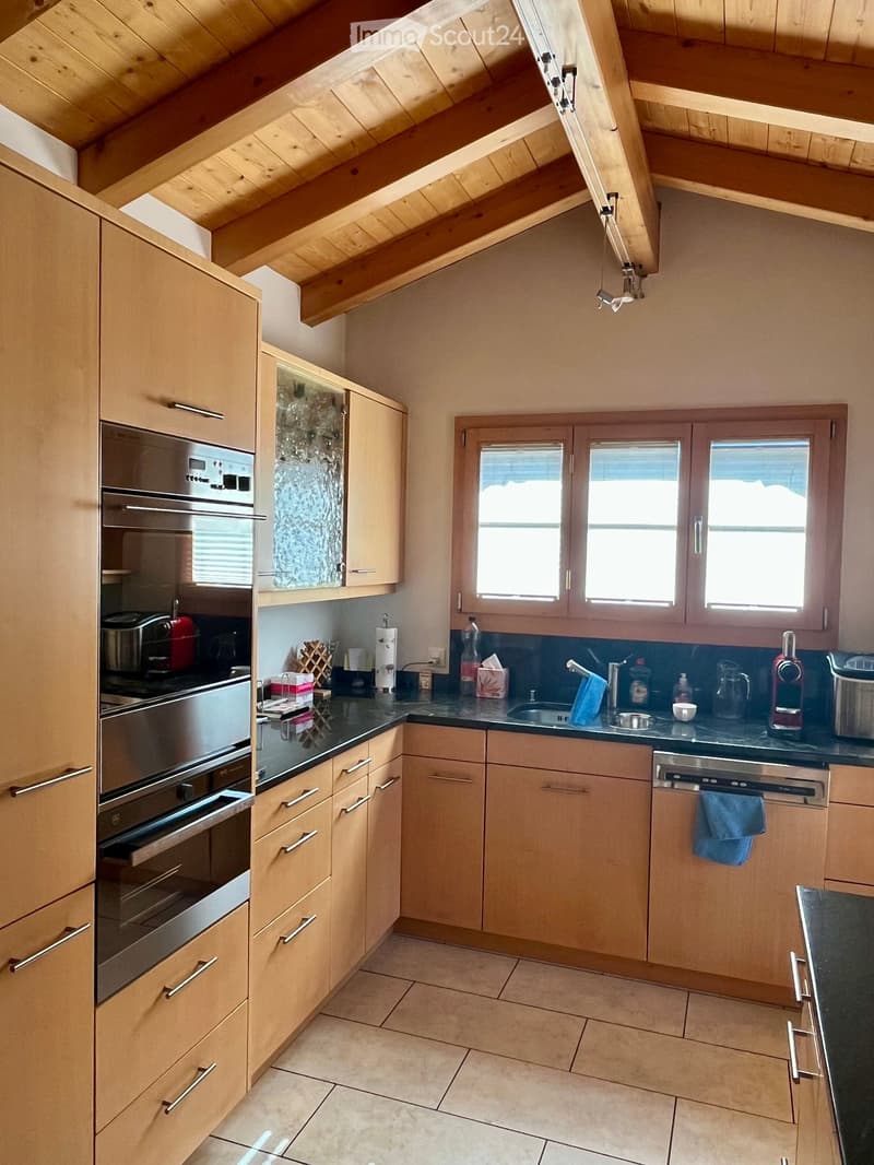 Modern kitchen with light wooden cabinets, stainless steel appliances, and a black countertop. Features include an oven, dishwasher, and various small appliances on the counter.