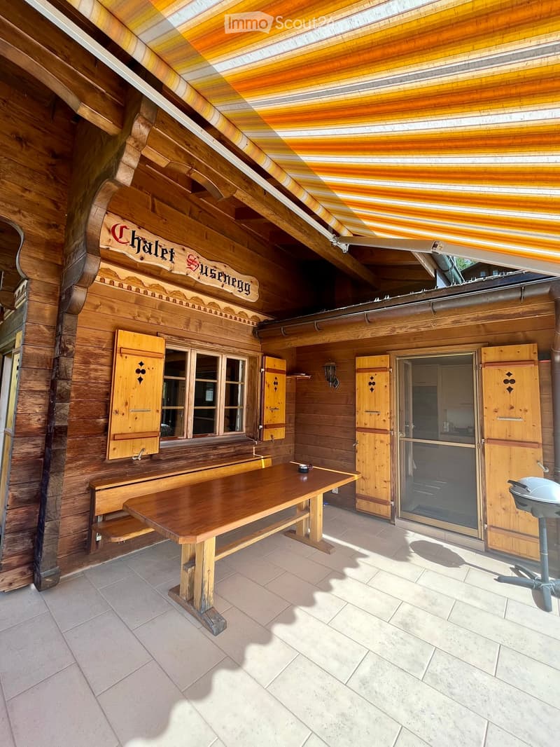 Wooden outdoor area with a large table, bench, and grill, featuring wooden walls and a slanted roof with striped awning.