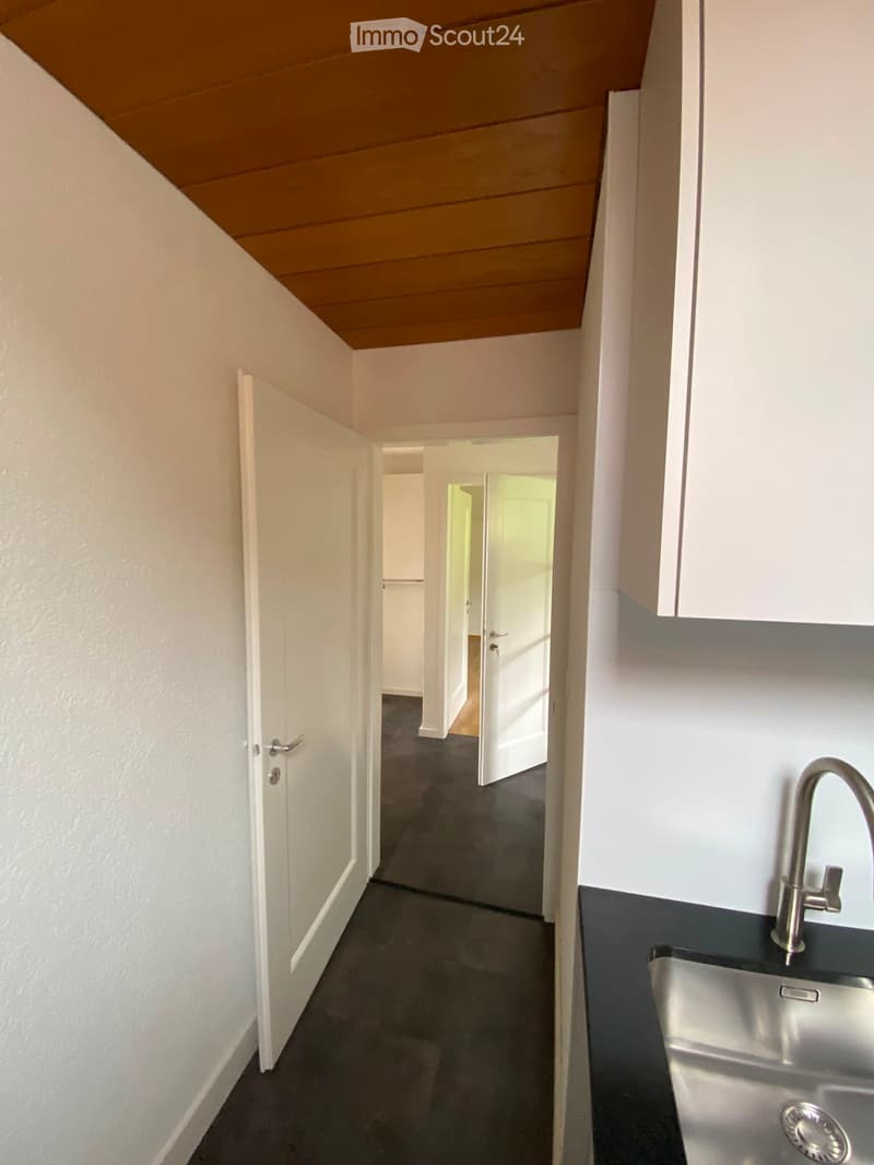 A narrow hallway with white walls, dark flooring, and several open doors leading to other rooms. A modern kitchen area is visible on the right with a stainless steel sink and faucet.