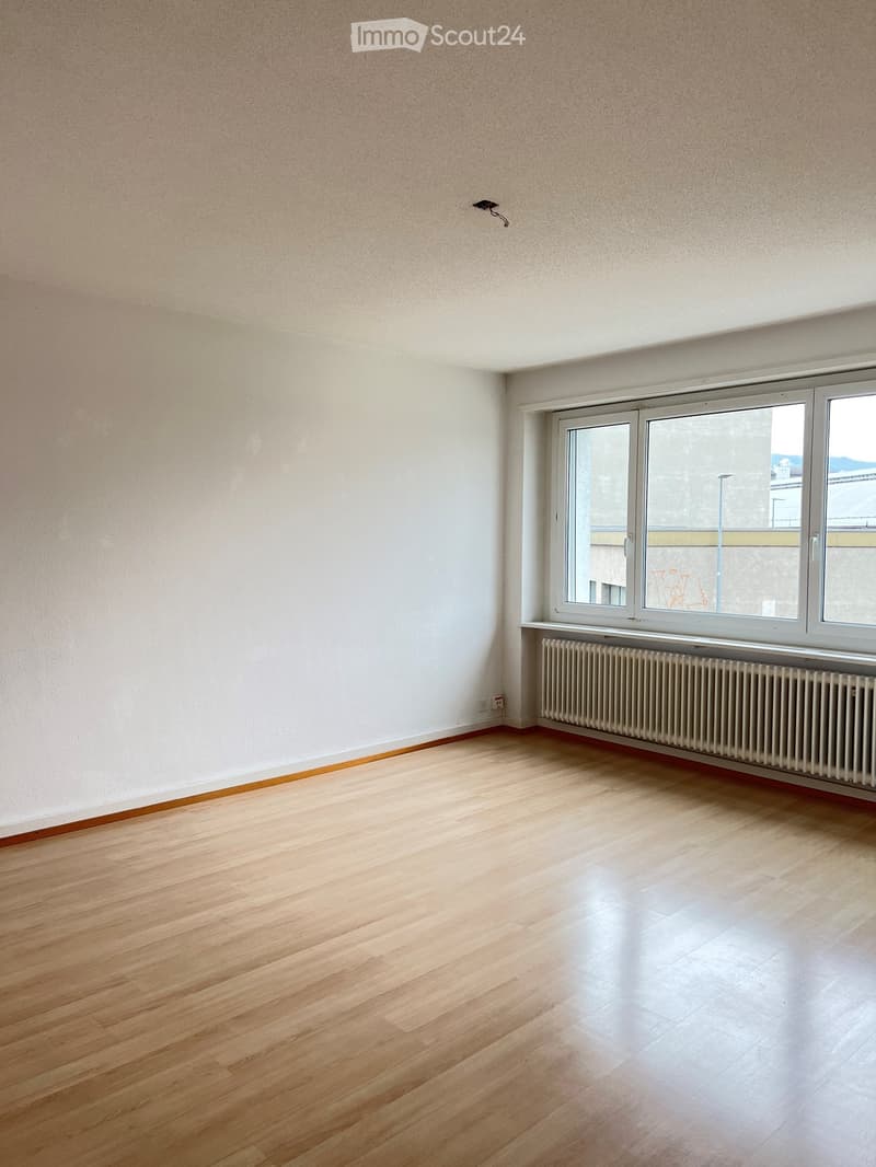 Empty room with light wooden flooring, white walls, and large windows with a radiator below them.