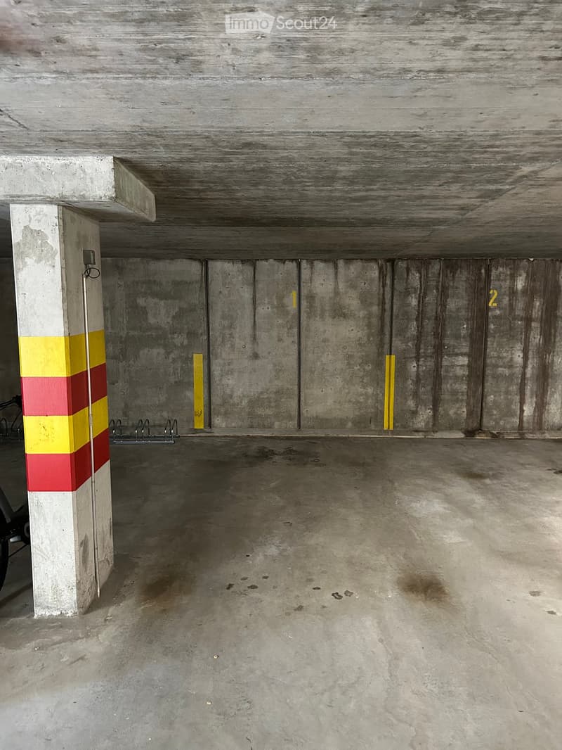 Empty underground parking space with concrete walls, floor, and ceiling. Features numbered parking spots and a single support column with a striped pattern.