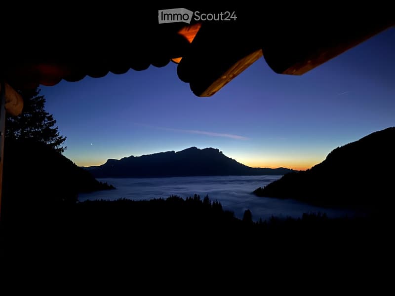 Balcony view at dusk showing a serene lake surrounded by mountains, with a clear sky and silhouettes of trees and structures.