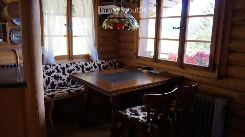 Cozy wooden room with large windows, a cowhide-patterned sofa, a wooden table, and a matching chair. A stained-glass lamp hangs from the ceiling.