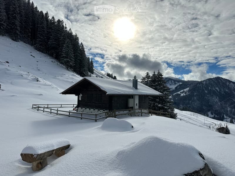 A wooden cabin with a snow-covered roof and a wooden deck, situated on a snowy slope surrounded by trees and mountains.
