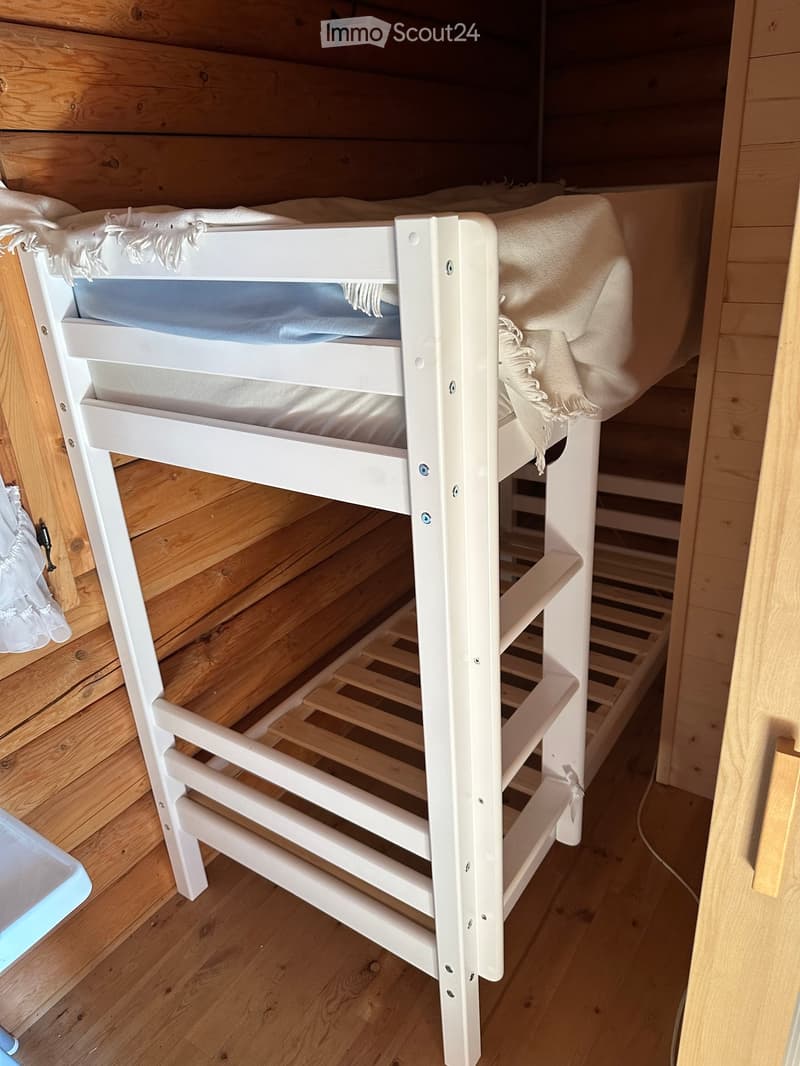 A white bunk bed with two mattresses and blankets, positioned against wooden walls and a wooden floor.