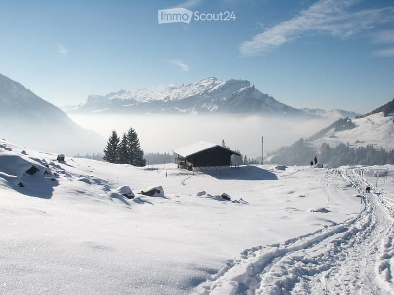 A snow-covered mountain landscape with a small wooden cabin, surrounded by snow-covered trees and mountains under a clear blue sky.