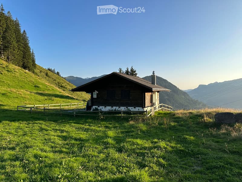 A wooden house with a dark roof and chimney, situated on a grassy slope surrounded by trees and mountains.
