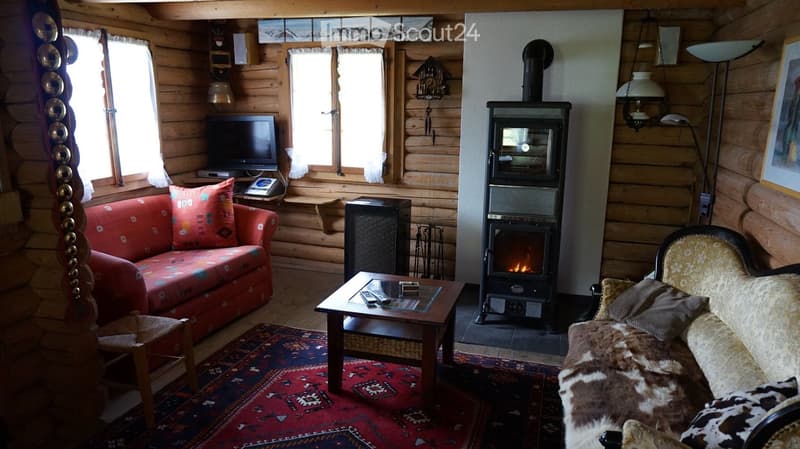 Cozy living room with wooden walls, a wood stove, a red patterned sofa, a coffee table, and a television. Features a large window with white curtains and a floor lamp.