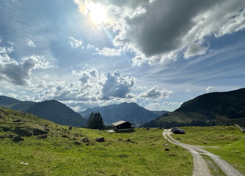 A scenic mountain valley with a small wooden cabin, surrounded by lush green grass, scattered rocks, and distant mountain peaks under a partly cloudy sky.