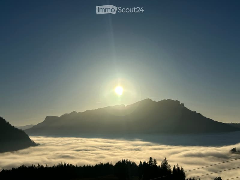 A serene mountain landscape with a clear blue sky, the sun shining brightly, and a layer of clouds below the peaks.