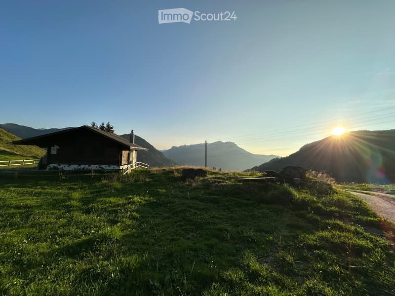 A wooden house sits on a grassy hill with a scenic mountain backdrop, surrounded by a peaceful landscape with a bench and a pathway.