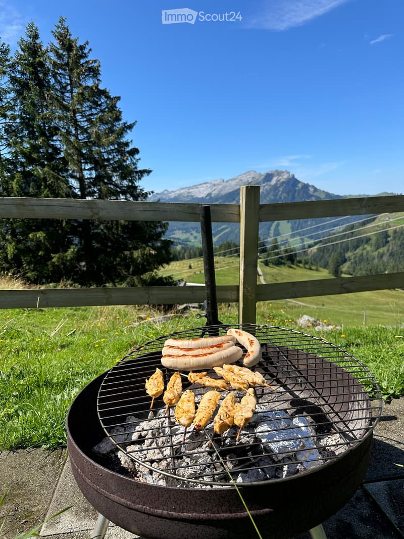 A barbecue grill with sausages and chicken skewers on a wooden deck, overlooking a scenic mountain landscape with green fields and trees.