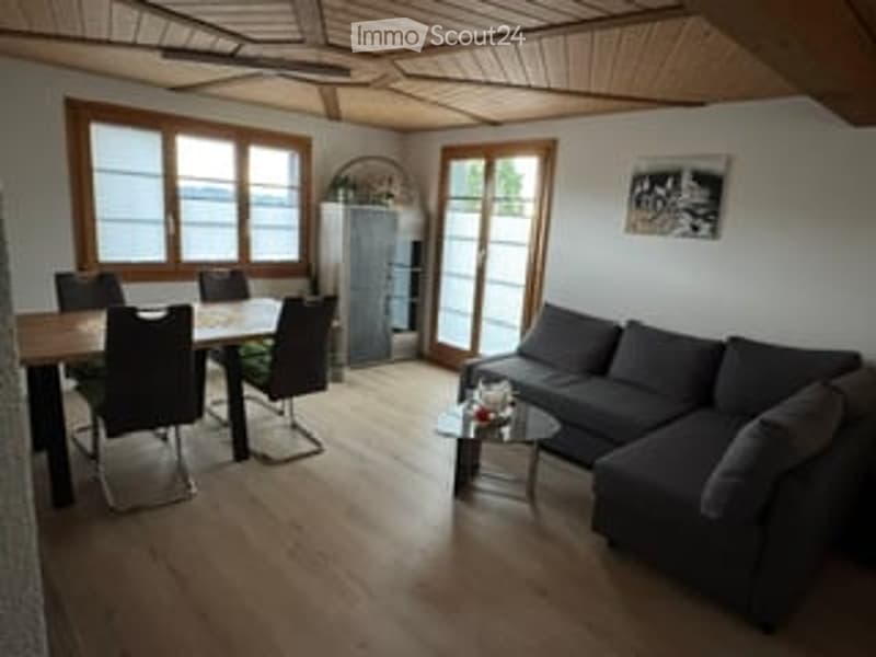 Bright living and dining area with wooden floors, large windows, a grey sectional sofa, a coffee table, a dining table with chairs, and a small cabinet.