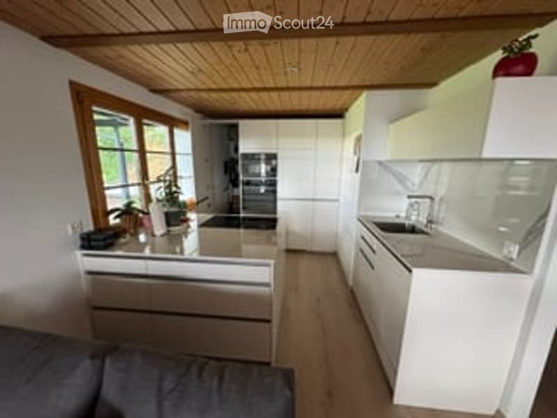 Modern kitchen with white cabinets, light wood flooring, and a central island with a sink and faucet. A potted plant is placed on the counter, and a refrigerator is visible in the background.