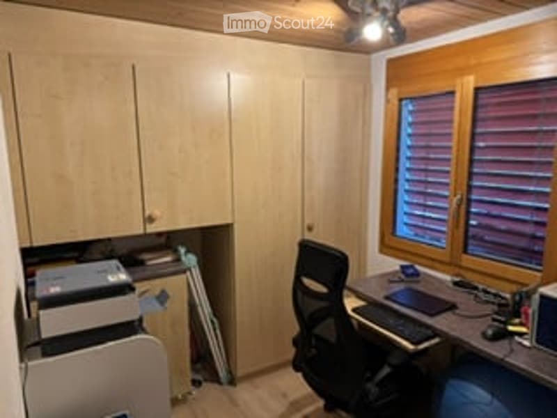 A small, well-lit room with wooden walls and ceiling, featuring a desk with a laptop, a chair, and a printer. There is a cabinet on the left side and a window with blinds on the right.