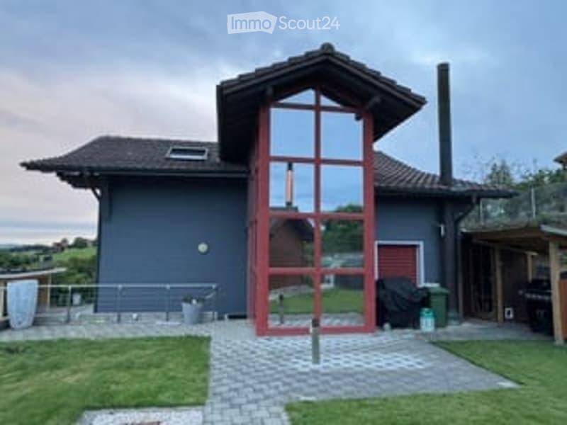 Modern two-story house with dark blue exterior, large red-framed glass entrance, and a small patio area with a grill and outdoor furniture.