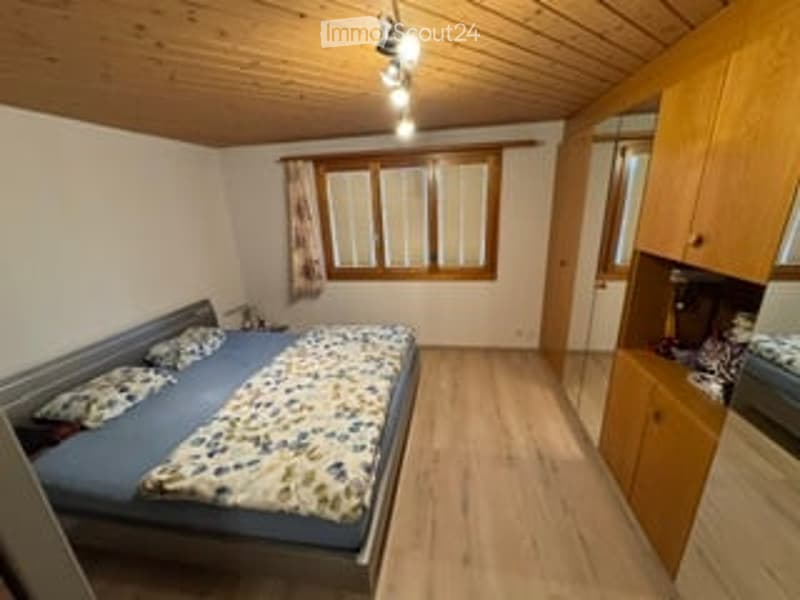 Cozy bedroom with wooden flooring, slanted wooden ceiling, large window with curtains, single bed with patterned bedding, nightstand, and built-in wardrobe with mirror.