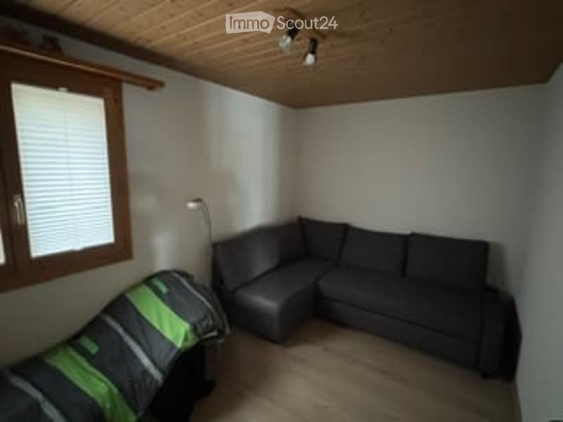 A small, modern living room with a grey sectional sofa, a wooden floor, and a large window with blinds. A floor lamp is positioned near the sofa.