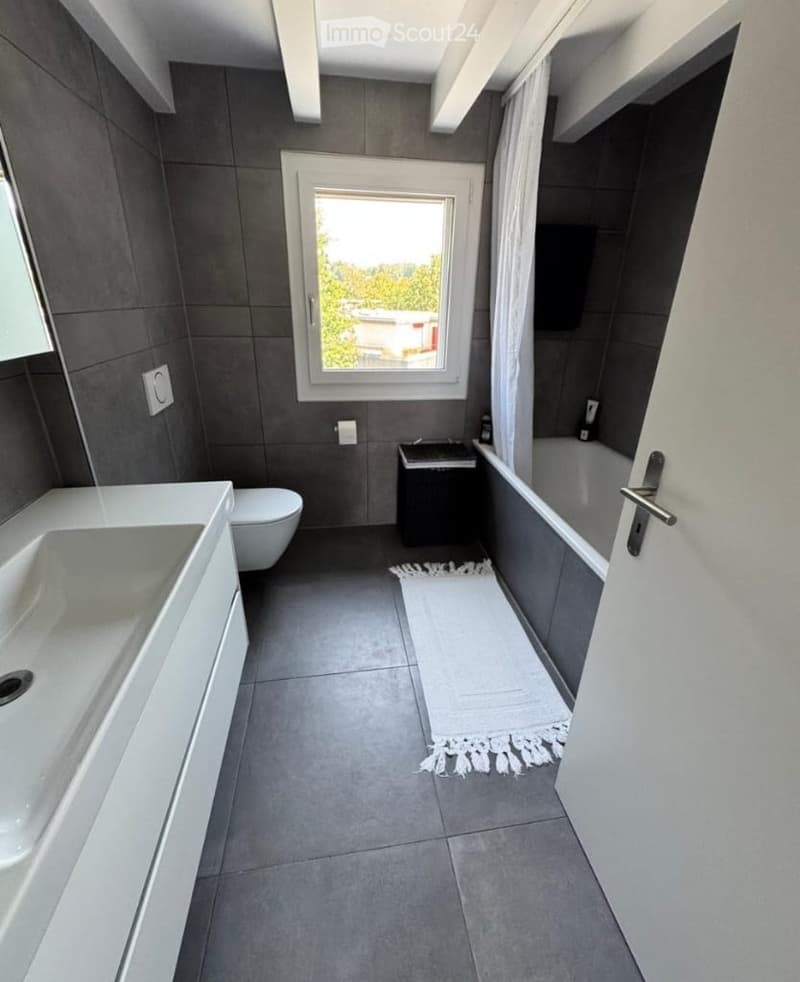 Modern bathroom with grey tiles, white sink, toilet, bathtub, and a small cabinet. Features a window with a view of greenery and a white rug on the floor.
