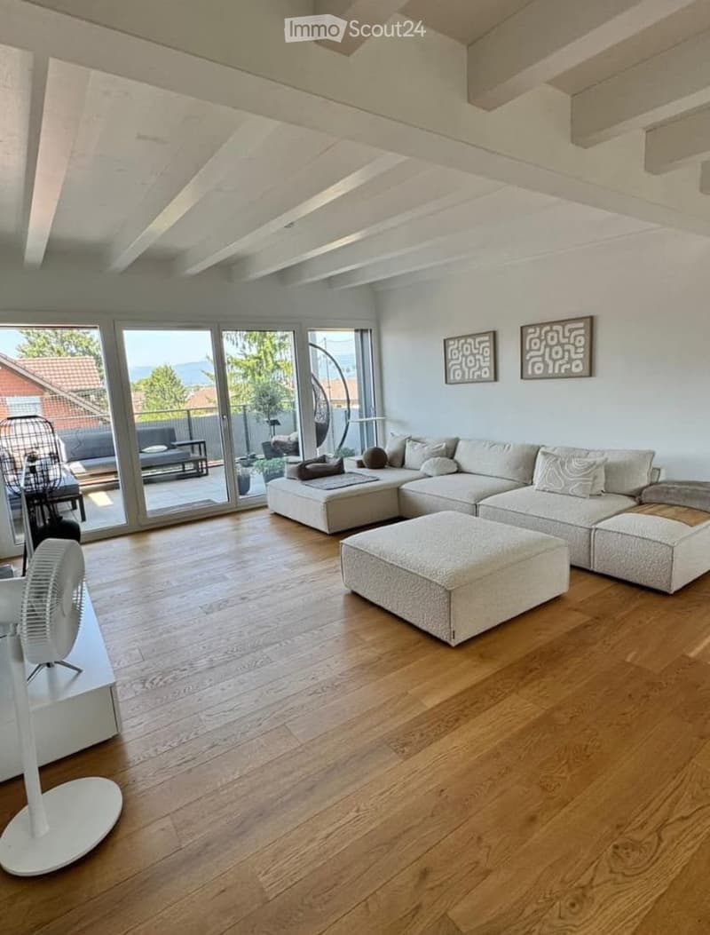 Modern living room with large windows offering a scenic view, featuring a sectional sofa, ottoman, and a white fan near the entrance.