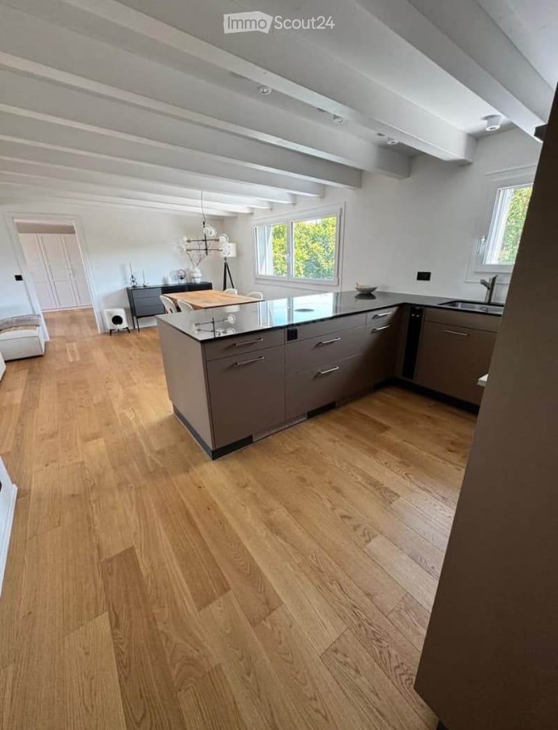 Modern kitchen with light wooden flooring, grey cabinets, central island, and pendant lighting. Visible elements include a sink, faucet, dining area with chairs and table, and a lamp.