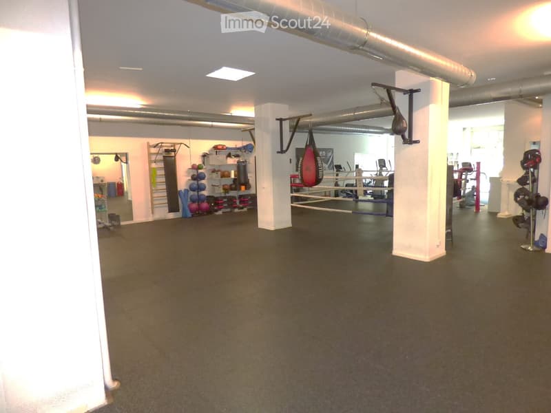 Well-lit gym space with boxing ring, punching bag, weight shelves, mirrors, and exercise bikes.