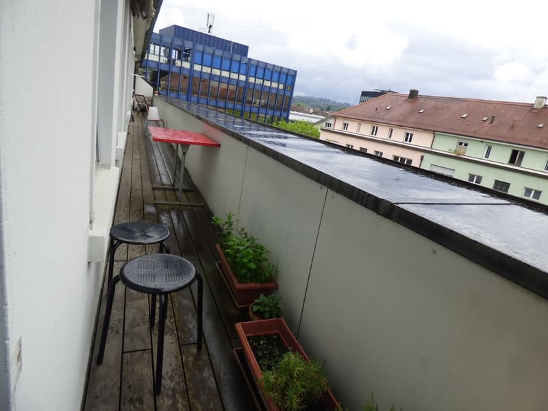 Balcony with wooden deck, two stools, a small table, and potted plants.