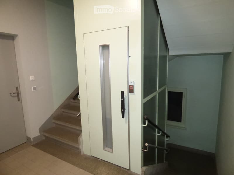 Hallway with a staircase, elevator door, and glass-paneled door leading to another area. Light-colored walls and tiled floor.