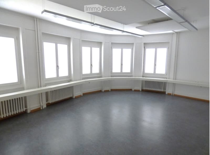 Empty room with white walls, large windows, grey carpeted floor, and radiators along the walls.