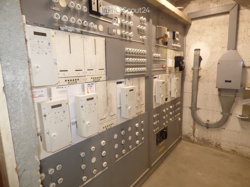 Electrical panel room with multiple circuit breakers, switches, and meters mounted on a grey wall. Concrete floor and exposed pipes visible.