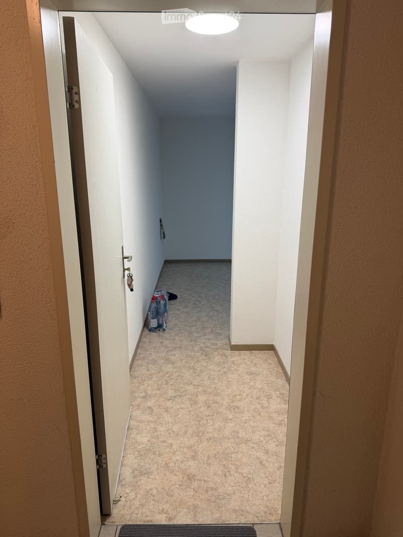 Empty hallway with beige carpet, white walls, and a single round ceiling light. A door is open on the left, and a small pile of water bottles is visible on the floor near the wall.