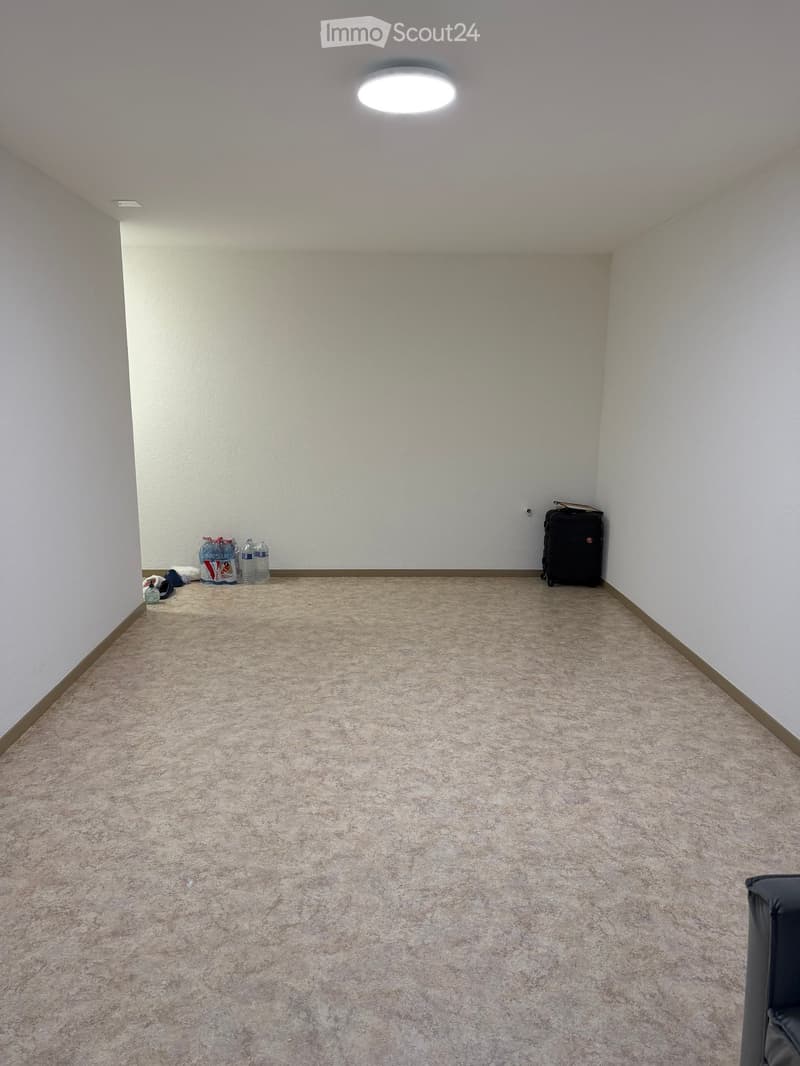 Empty room with beige carpet and white walls, featuring a ceiling light and a few scattered items including a suitcase and water bottles.