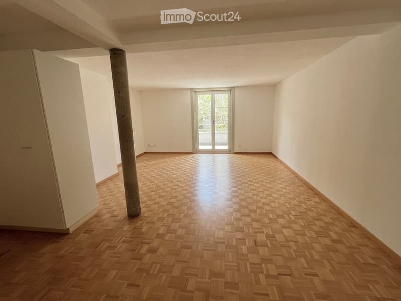 Empty room with wooden herringbone flooring, white walls, and a large window leading to a balcony.