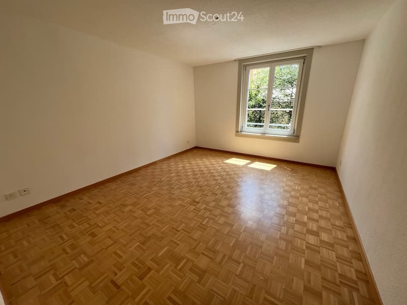 Empty room with wooden parquet flooring and a single window with white frame, allowing natural light to enter.