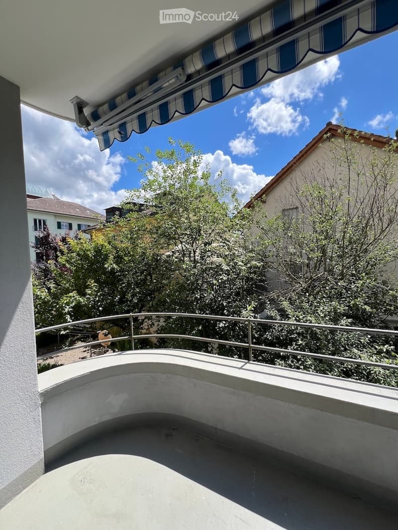 Balcony with a striped awning, metal railing, and view of trees and neighboring houses.