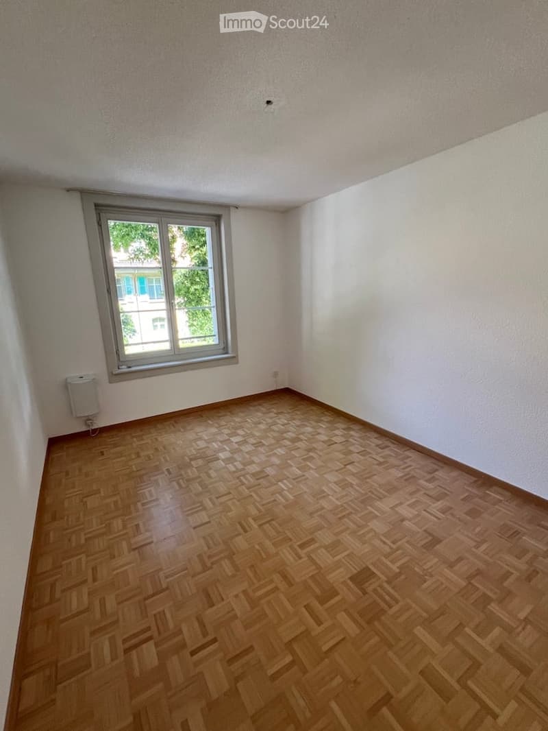 Empty room with wooden herringbone flooring, white walls, and a single window with a view of greenery outside.