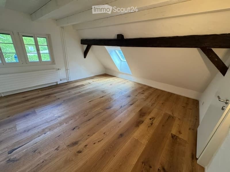 Empty attic room with wooden flooring, white walls, and exposed wooden beams. Three windows and a skylight provide natural light.