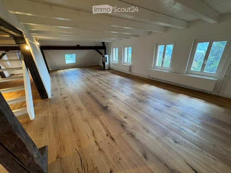Completely empty room with wooden floor and exposed wooden beams on the ceiling. Natural light from multiple windows.