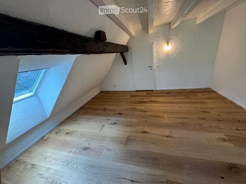 Empty attic room with wooden flooring, white walls, skylight window, and exposed wooden beams on the ceiling.