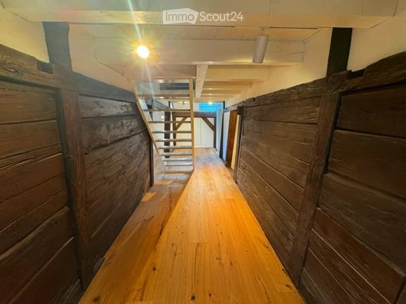 A narrow wooden hallway with exposed wooden walls and a wooden staircase leading upwards, illuminated by a single overhead light.