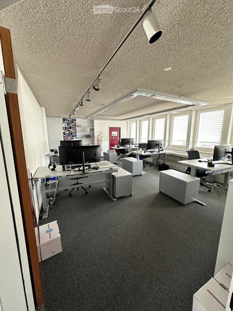 Office space with multiple desks, chairs, computers, and a large window providing natural light. The room features a carpeted floor and modern lighting fixtures.