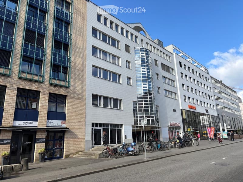 Modern multi-story building with large windows and a glass tower section, located on a street with bicycles parked in front.